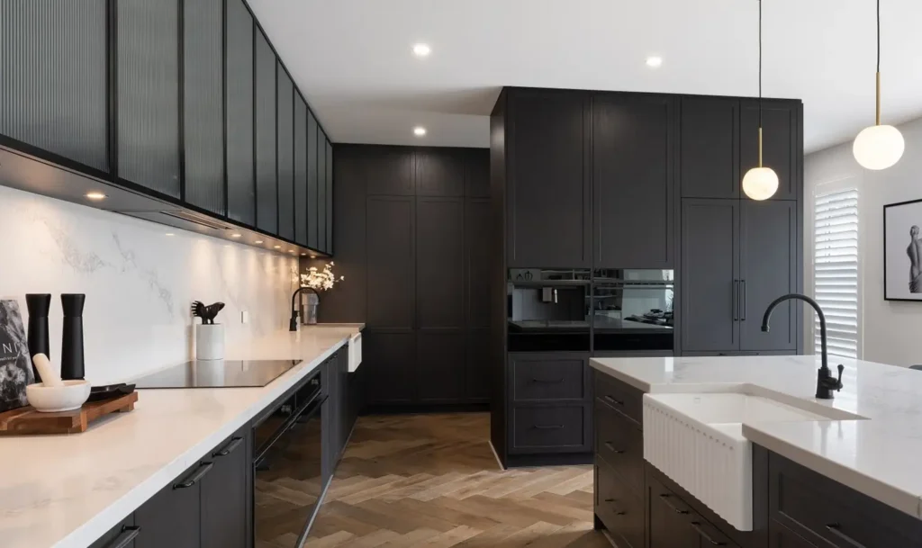 Black kitchen with white marble countertops, farmhouse sink, black faucet, globe pendant lights, and herringbone wood floor.